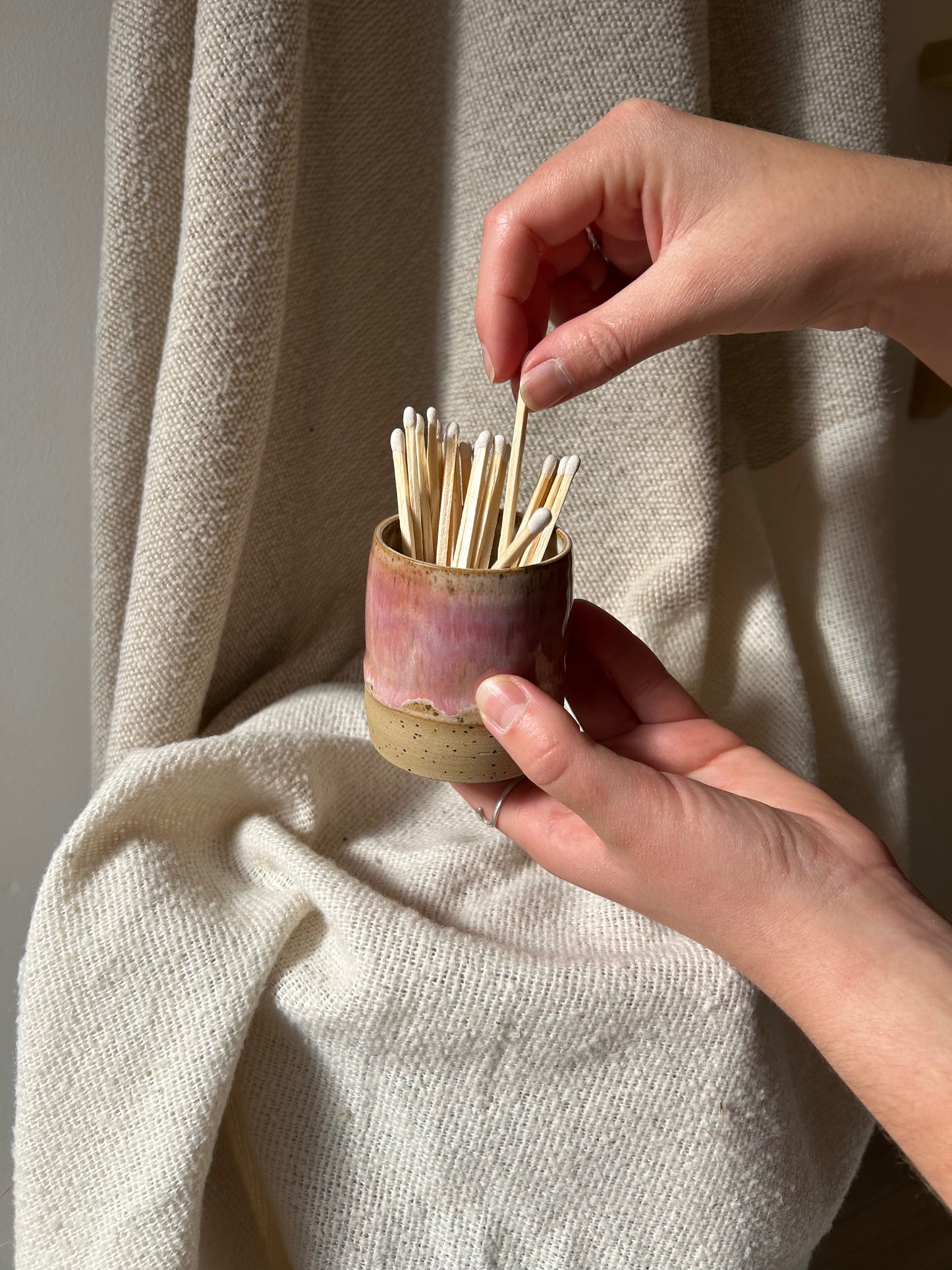 A person's hands holding a small, rustic ceramic matchstick container filled with wooden matchsticks, set against a draped beige linen background.