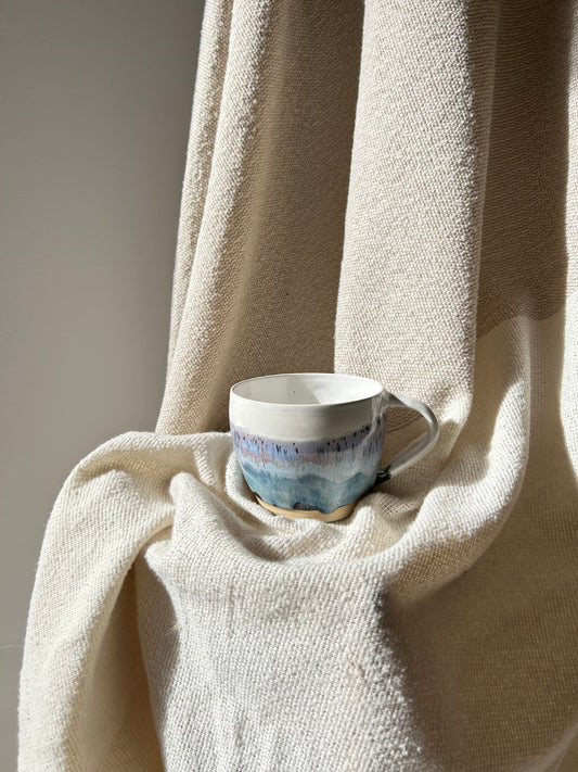 A ceramic coffee mug with a blue and white abstract landscape design, resting on draped cream-colored linen fabric.