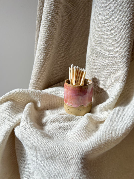 A small cylindrical ceramic holder with a pink and beige marbled finish, filled with wooden matchsticks, set against draped cream-colored woven fabric.