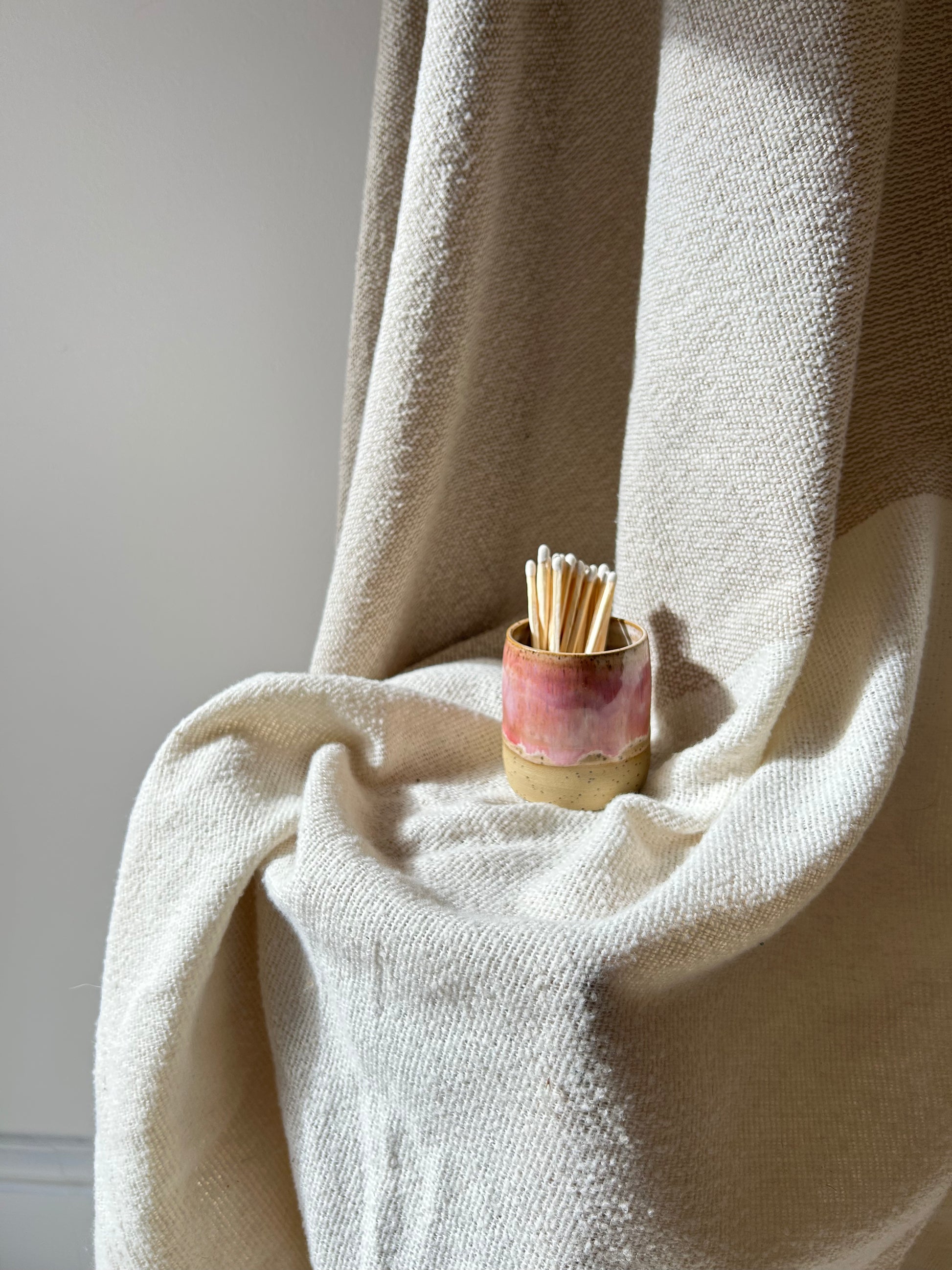 A small handmade ceramic holder filled with wooden matchsticks, placed on draped cream-colored fabric against a neutral wall.