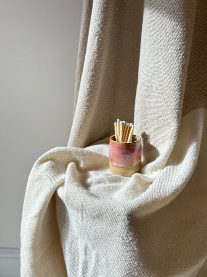 A small handmade ceramic holder filled with wooden matchsticks, placed on draped cream-colored fabric against a neutral wall.