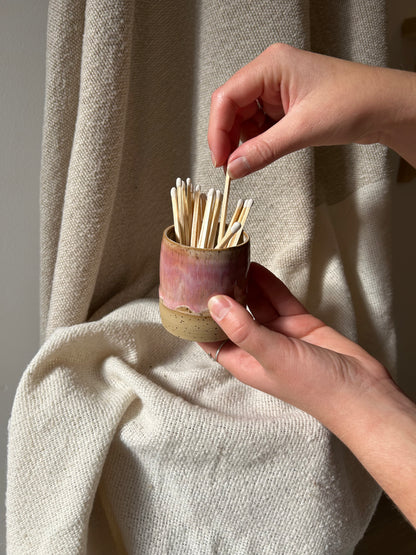 A person's hands holding a small, rustic ceramic matchstick container filled with wooden matchsticks, set against a draped beige linen background.