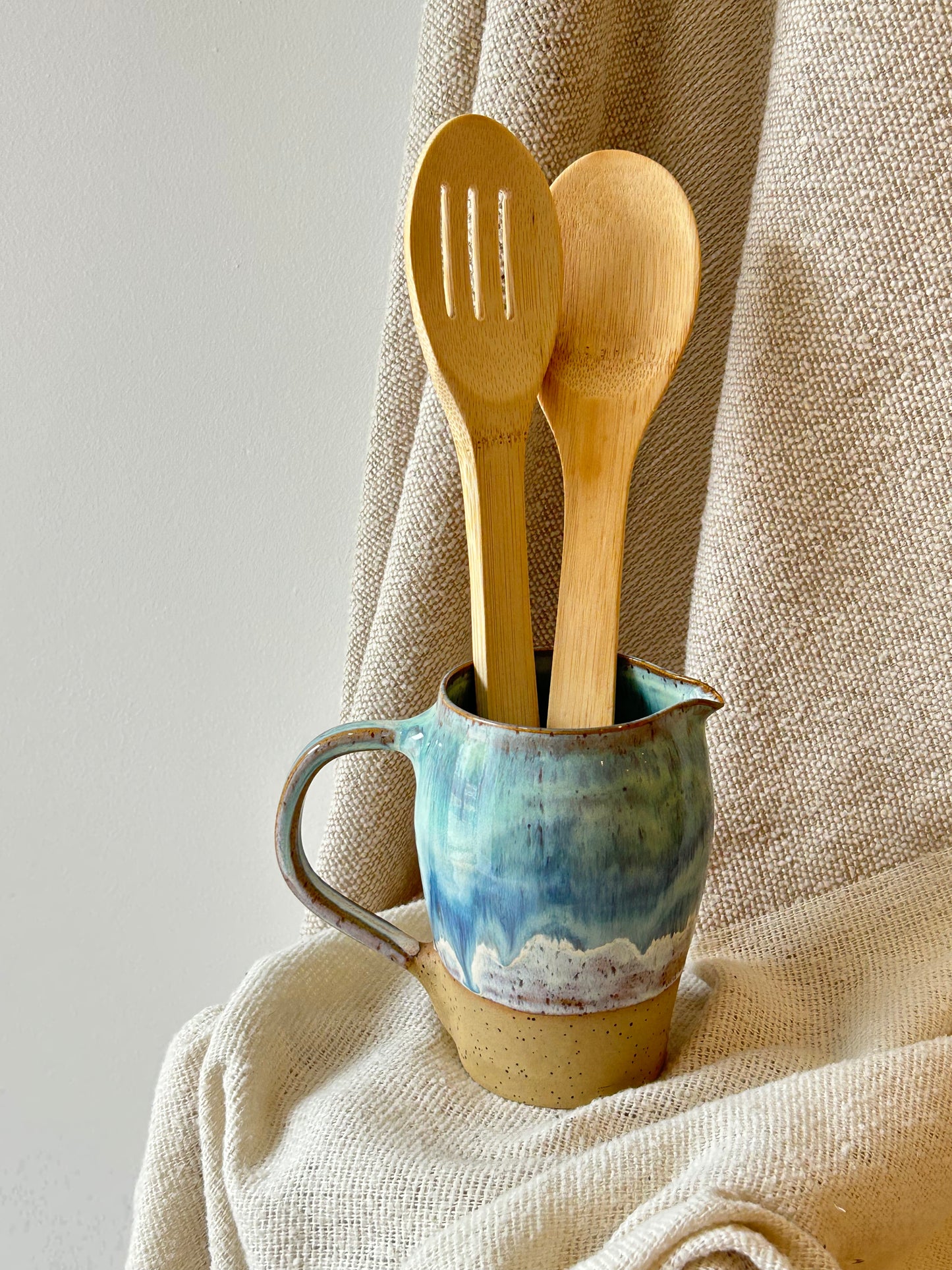 Two light natural wood kitchen utensils, a slotted spoon and a spoon, standing upright in a small glazed ceramic pitcher with blue, green, and beige tones, set against a draped beige linen fabric background.