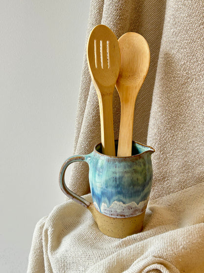 Two light natural wood kitchen utensils, a slotted spoon and a spoon, standing upright in a small glazed ceramic pitcher with blue, green, and beige tones, set against a draped beige linen fabric background.