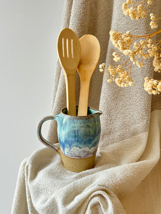Two light natural wood kitchen utensils, a slotted spoon and a wooden spoon, standing in a small blue and beige ceramic pitcher filled with water, set against a draped beige linen fabric backdrop with dried flowers.
