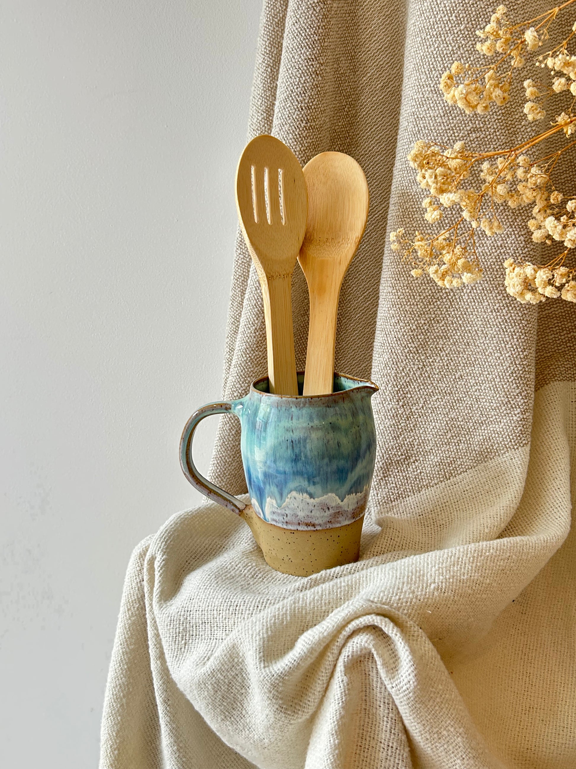 Two light natural wood kitchen utensils, one slotted spoon and one spoon, standing in a small blue and beige ceramic pitcher filled with water, set against a draped beige linen fabric backdrop with dried flowers.