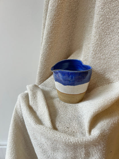 A small handmade ceramic cup with a deep blue glazed top, a white middle band, and a speckled beige clay base, resting on draped cream linen fabric.