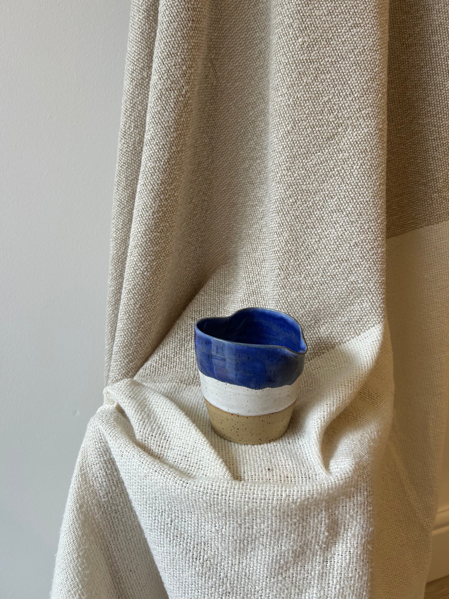 A handmade ceramic cup with a deep blue glazed top half and a natural beige clay bottom half, resting on draped cream-colored linen fabric.