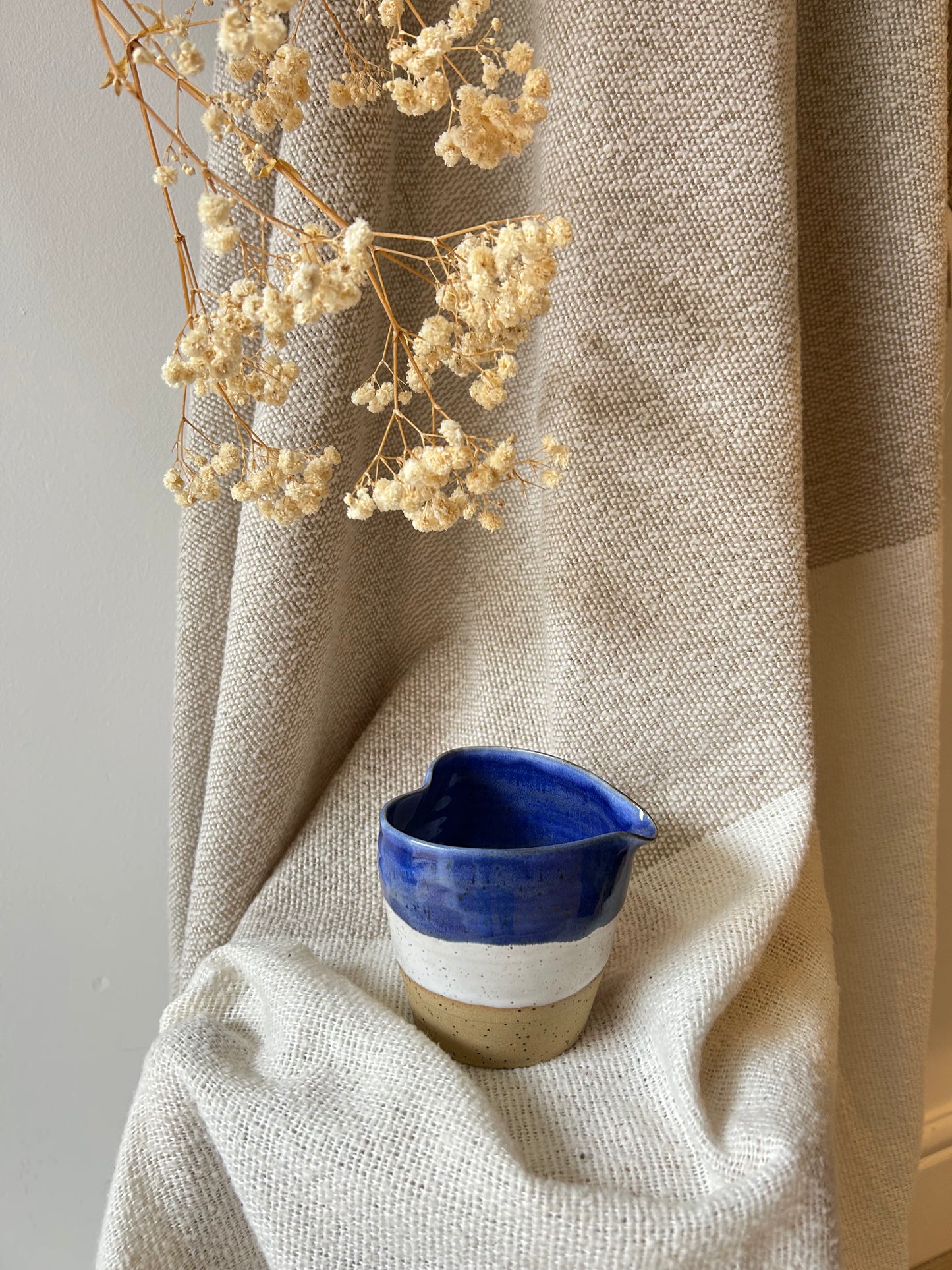 A small handmade ceramic pitcher with a blue and white glazed finish sits on draped beige linen fabric, with dried cream flowers hanging above in a styled interior setting.