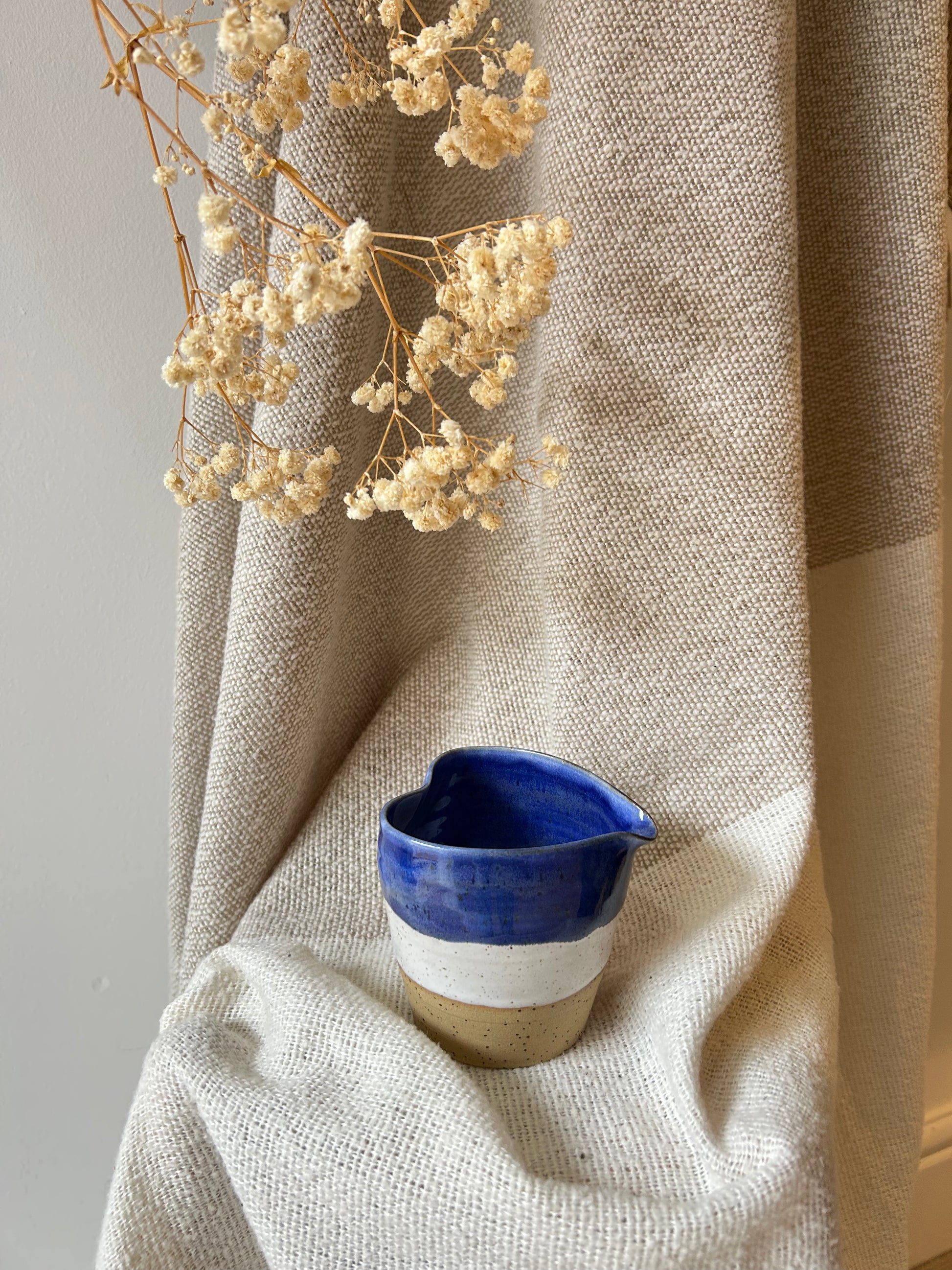 A small handmade ceramic pitcher with a blue and white glazed finish sits on draped beige linen fabric, with dried cream flowers hanging above in a styled interior setting.