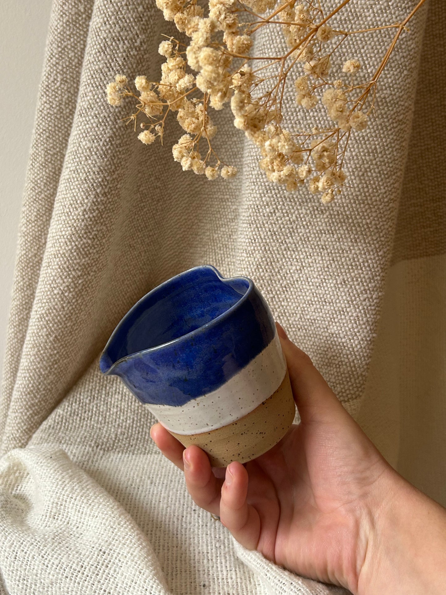 A hand holds a small handmade ceramic cup with a glossy deep blue top half, a white middle band, and a speckled beige lower half, set against a draped beige fabric backdrop with dried flowers.