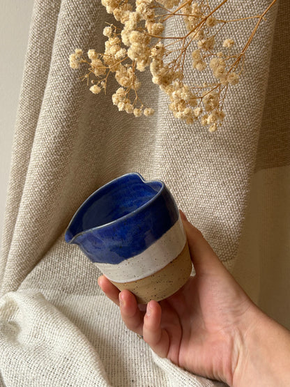 A hand holds a small handmade ceramic cup with a glossy deep blue top half, a white middle band, and a speckled beige lower half, set against a draped beige fabric backdrop with dried flowers.