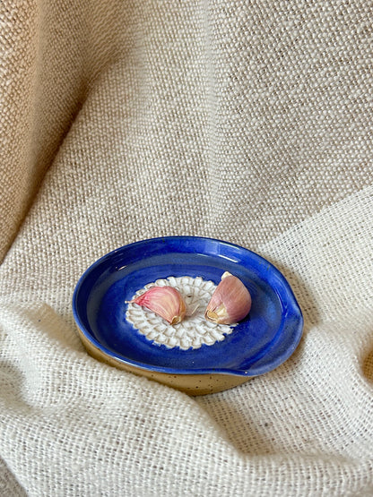A small oval ceramic garlic grater with a deep cobalt blue glazed top and a natural beige clay base, holding two garlic cloves on a white patterned coaster, set against a textured beige linen background.