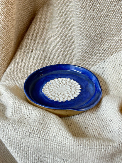 A small oval ceramic garlic grater with a deep cobalt blue glazed exterior and a white textured floral pattern in the center, resting on a beige linen fabric background.