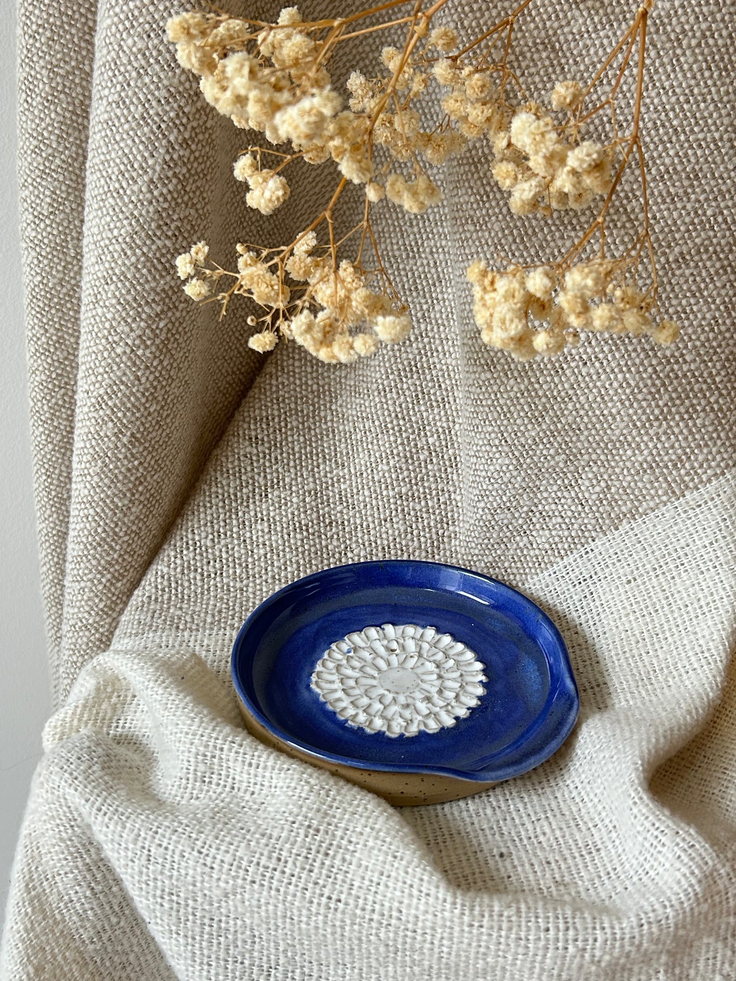 A small handmade ceramic garlic grater with a deep cobalt blue glazed exterior and a white textured floral pattern in the center, styled on a beige linen cloth with dried flowers.