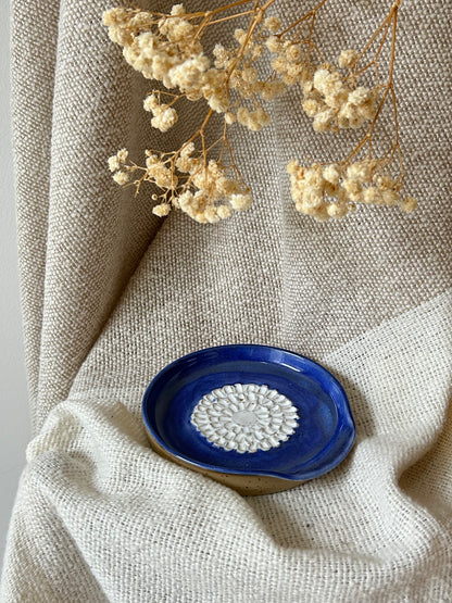 A small handmade ceramic garlic grater with a deep cobalt blue glazed exterior and a white textured floral pattern in the center, styled on a beige linen cloth with dried flowers.