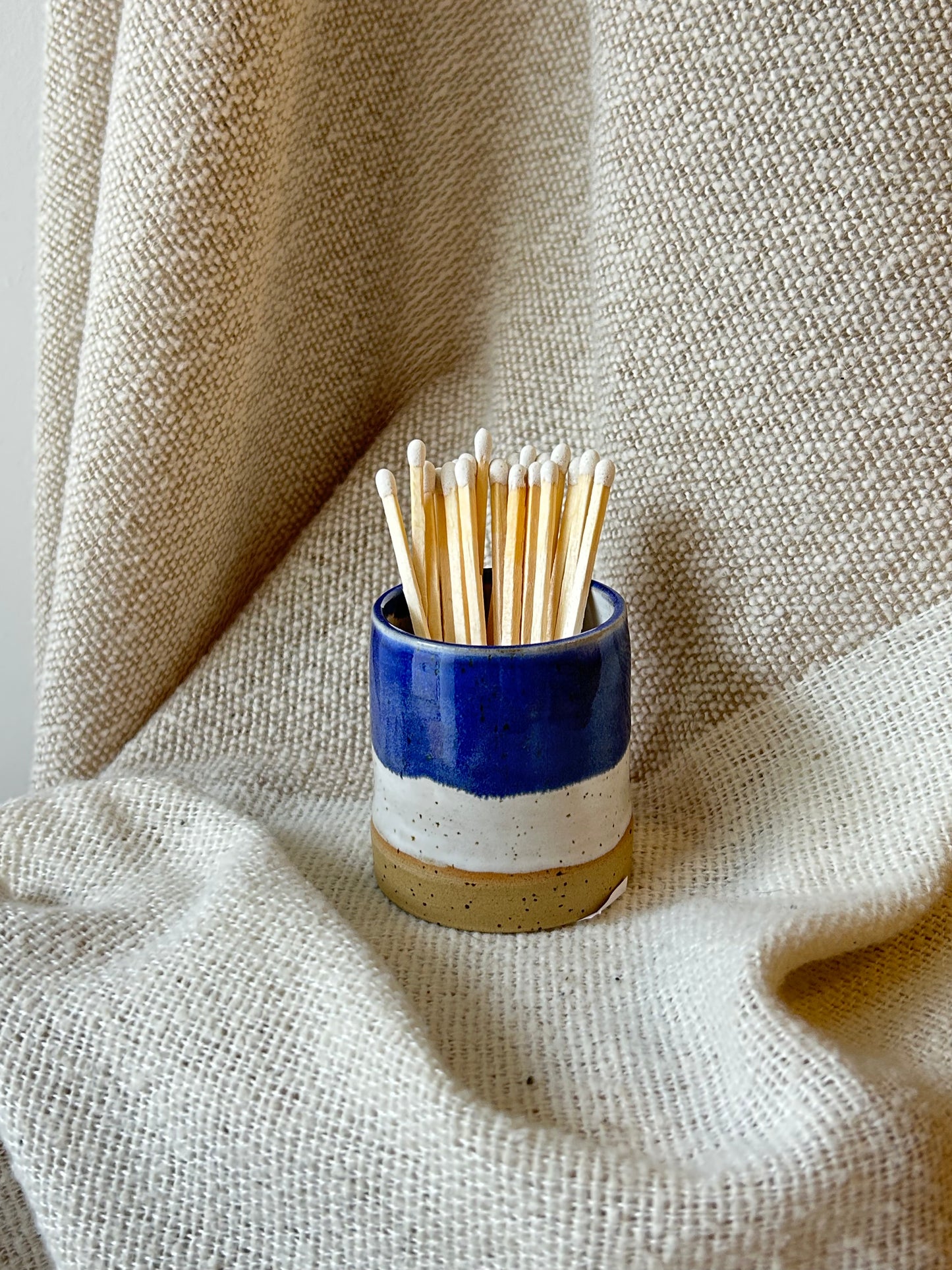 A small ceramic matchstick pot with a blue, white, and beige glaze, filled with wooden toothpicks, set against a draped beige linen fabric background.