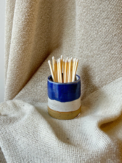 A small ceramic matchstick pot with a blue, white, and beige glaze, filled with wooden toothpicks, set against a draped beige linen fabric background.