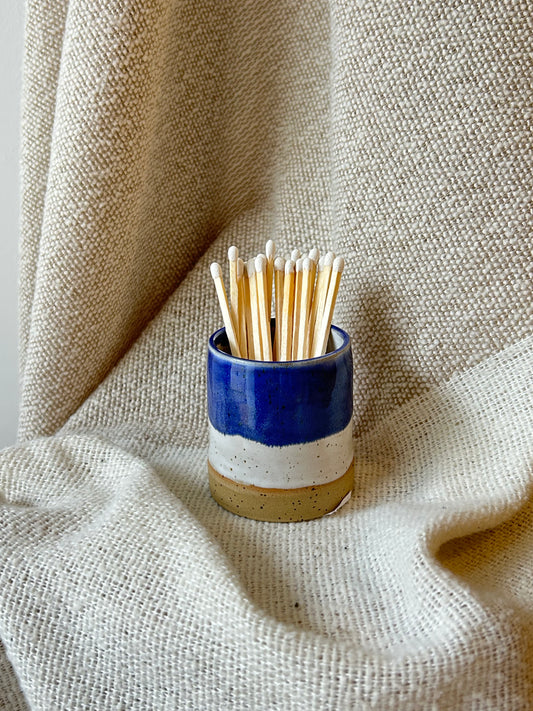 A small ceramic matchstick pot with a blue, white, and beige glaze, filled with wooden toothpicks, set against a draped beige linen fabric background.