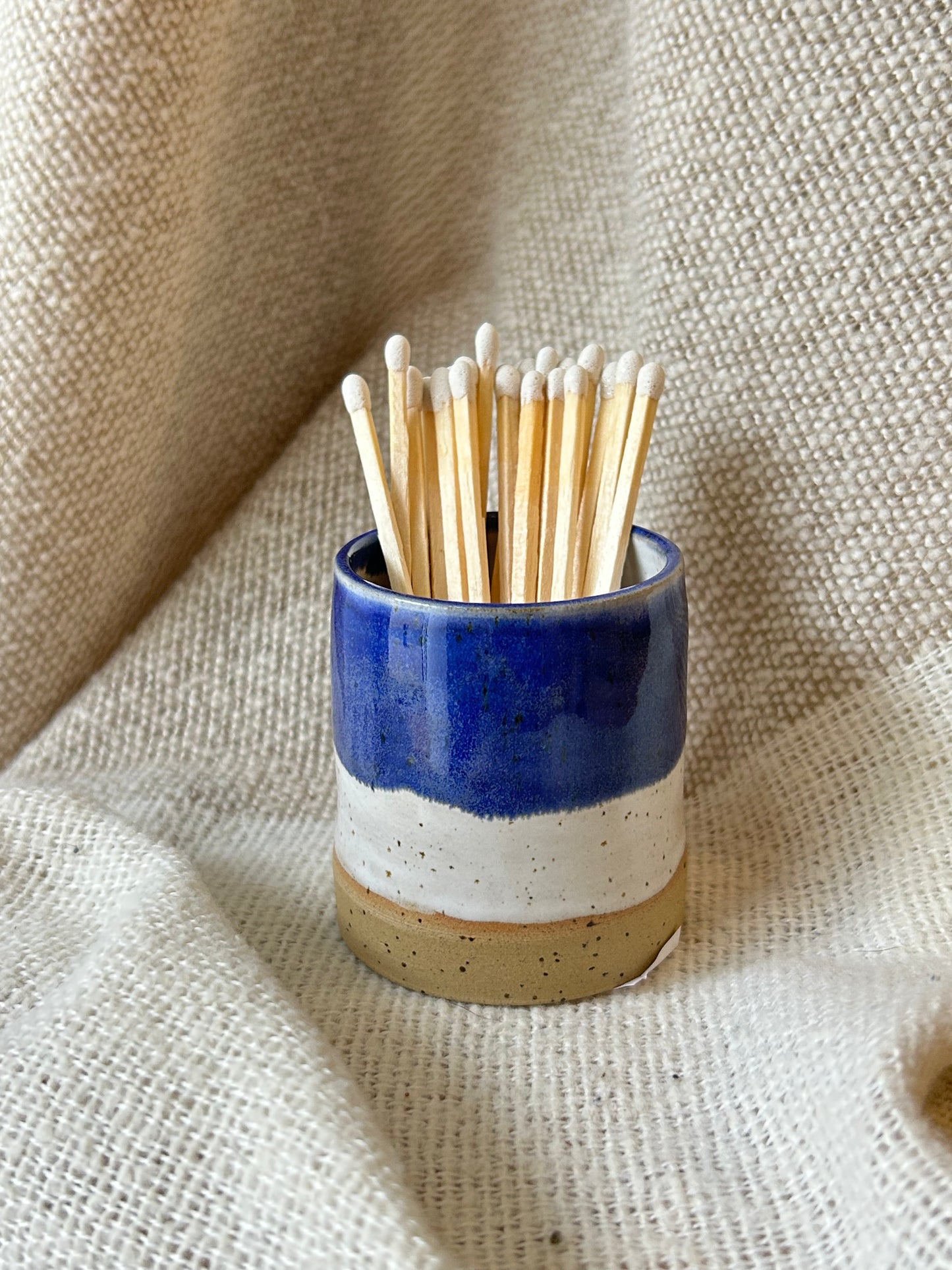A small ceramic match holder with a blue, white, and beige glaze, filled with wooden matchsticks, set against a draped beige linen fabric background.