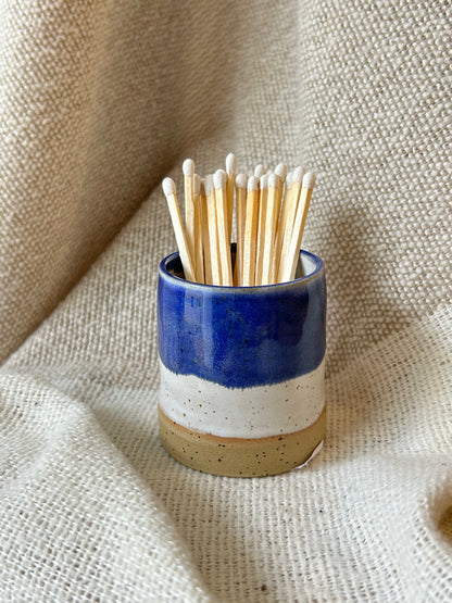 A small ceramic match holder with a blue, white, and beige glaze, filled with wooden matchsticks, set against a draped beige linen fabric background.