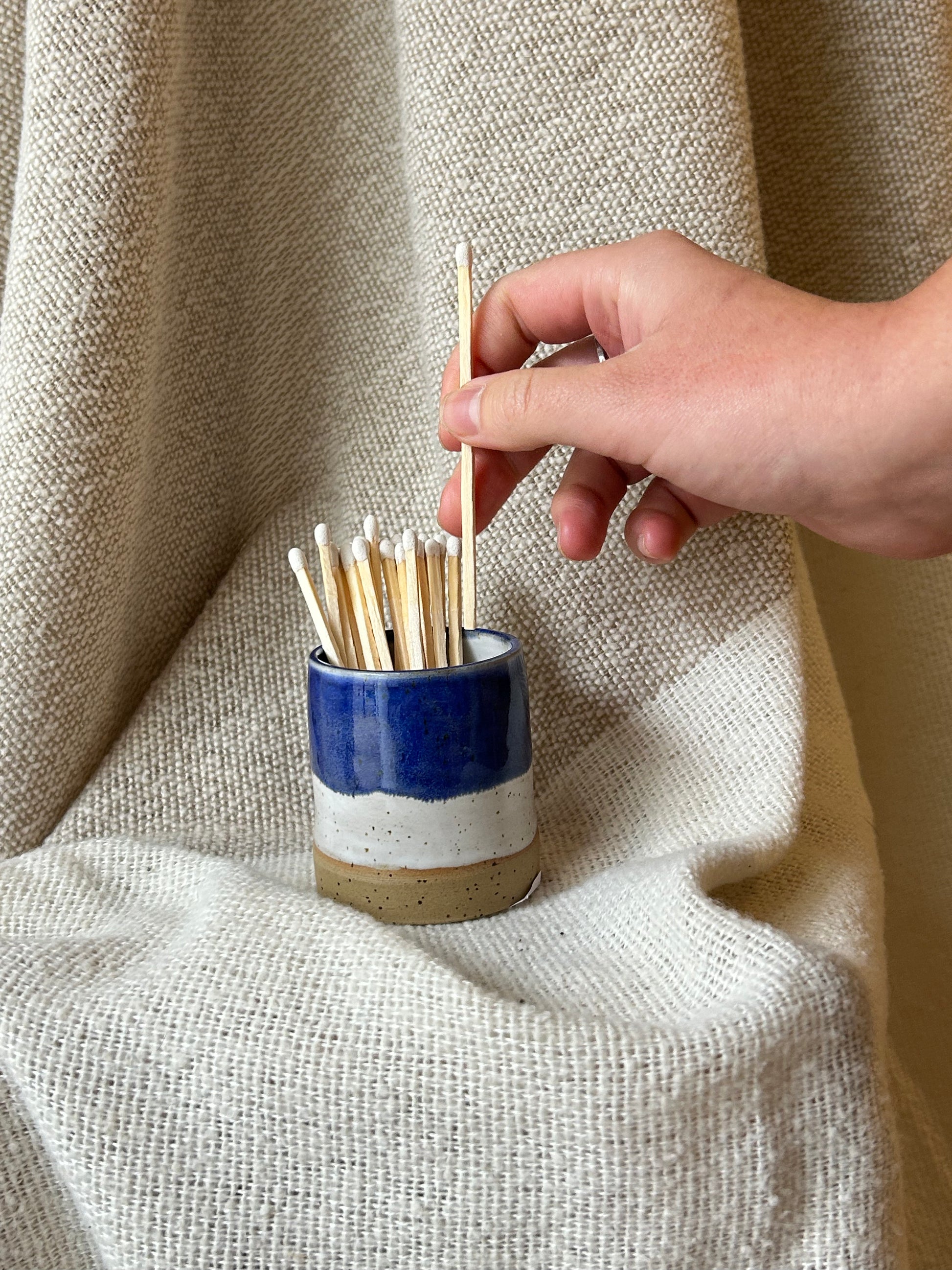 A hand holding a wooden matchstick above a small ceramic holder filled with matchsticks, set against a draped beige linen fabric background.
